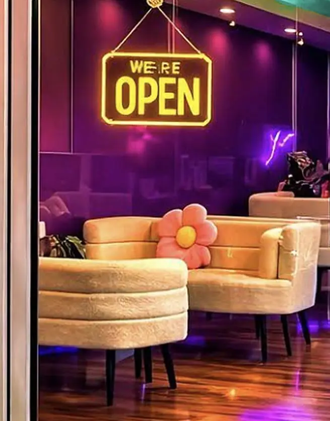Through the glass door: curved cream sofas and a pink flower cushion under violet light, beneath a glowing sign that reads We're open.